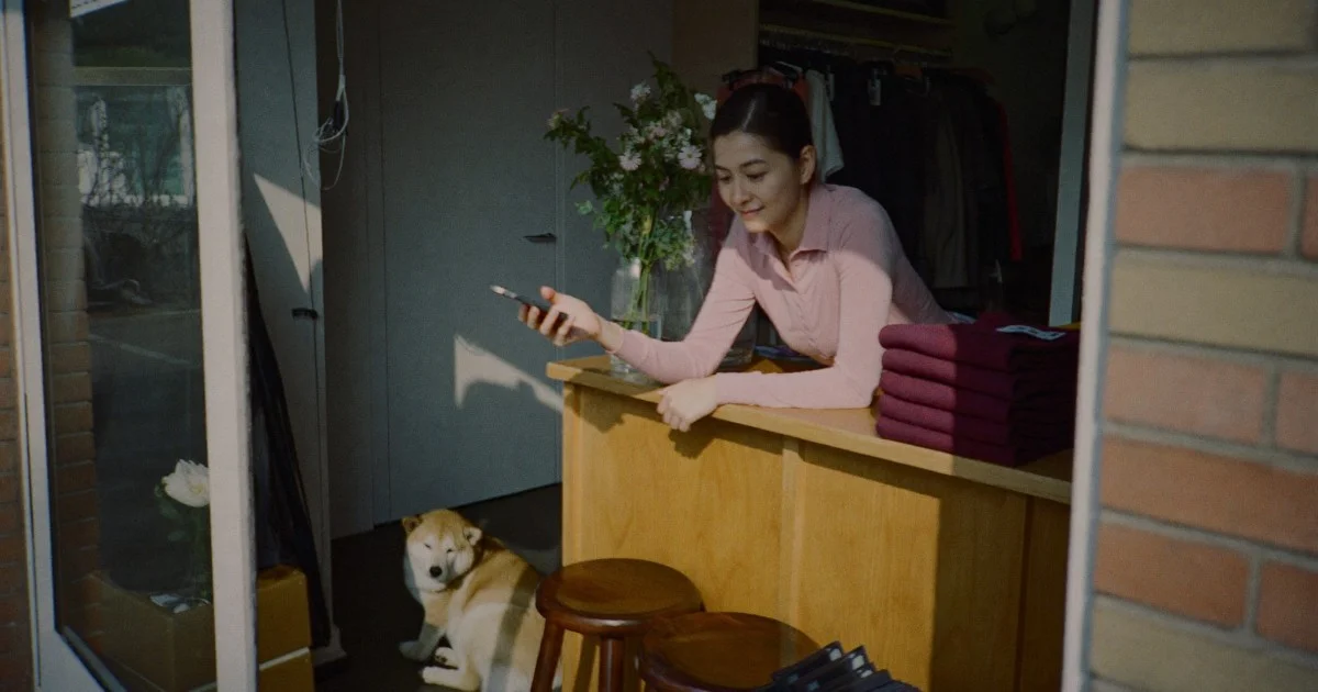 A boutique owner leaning on her shop counter scrolling her phone, a stack of burgundy knitwear beside her and a dog asleep on the floor.