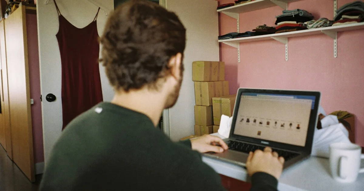 Over the shoulder of a man working on a laptop in a spare-room stockroom, shelves of folded garments and a burgundy dress on the wardrobe door.