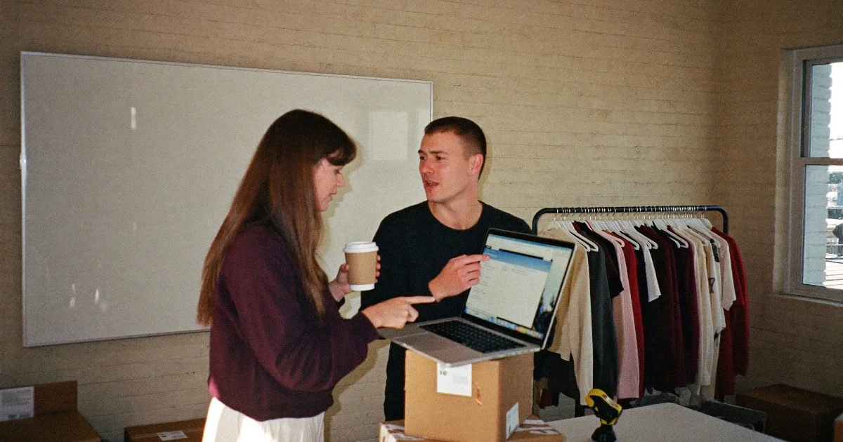 Two colleagues in a small e-commerce office reviewing results on a laptop balanced on shipping boxes, a rail of garments behind them.