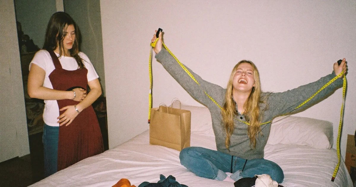 One friend holds a burgundy dress against herself while the other sits on the bed laughing with a tape measure stretched between her hands.