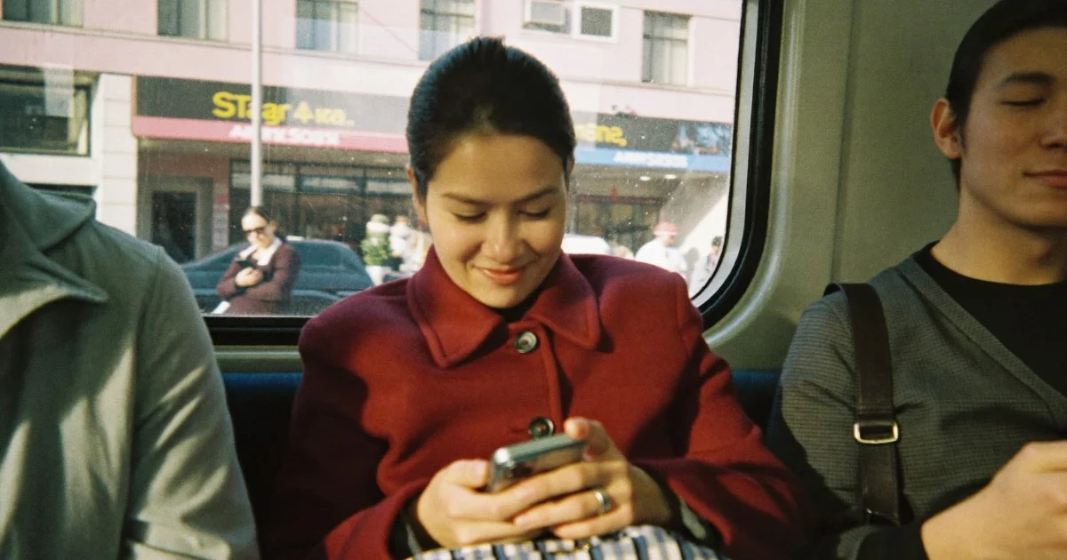A woman in a burgundy wool coat on a tram, smiling down at her phone in late afternoon light.